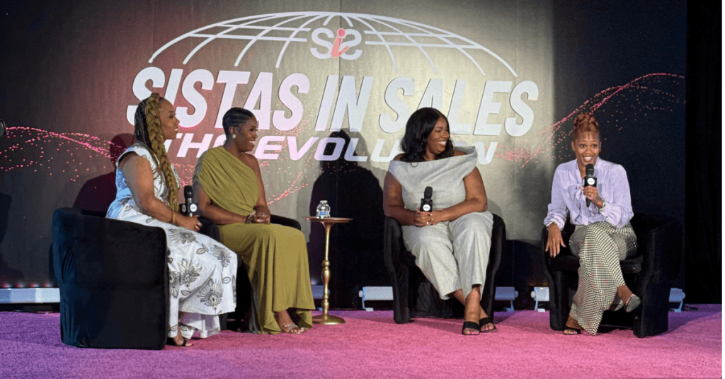 Four women seated on stage at the 'Sistas in Sales' event, discussing topics related to sales and empowerment. The backdrop features the event logo, and the setting has a vibrant atmosphere.